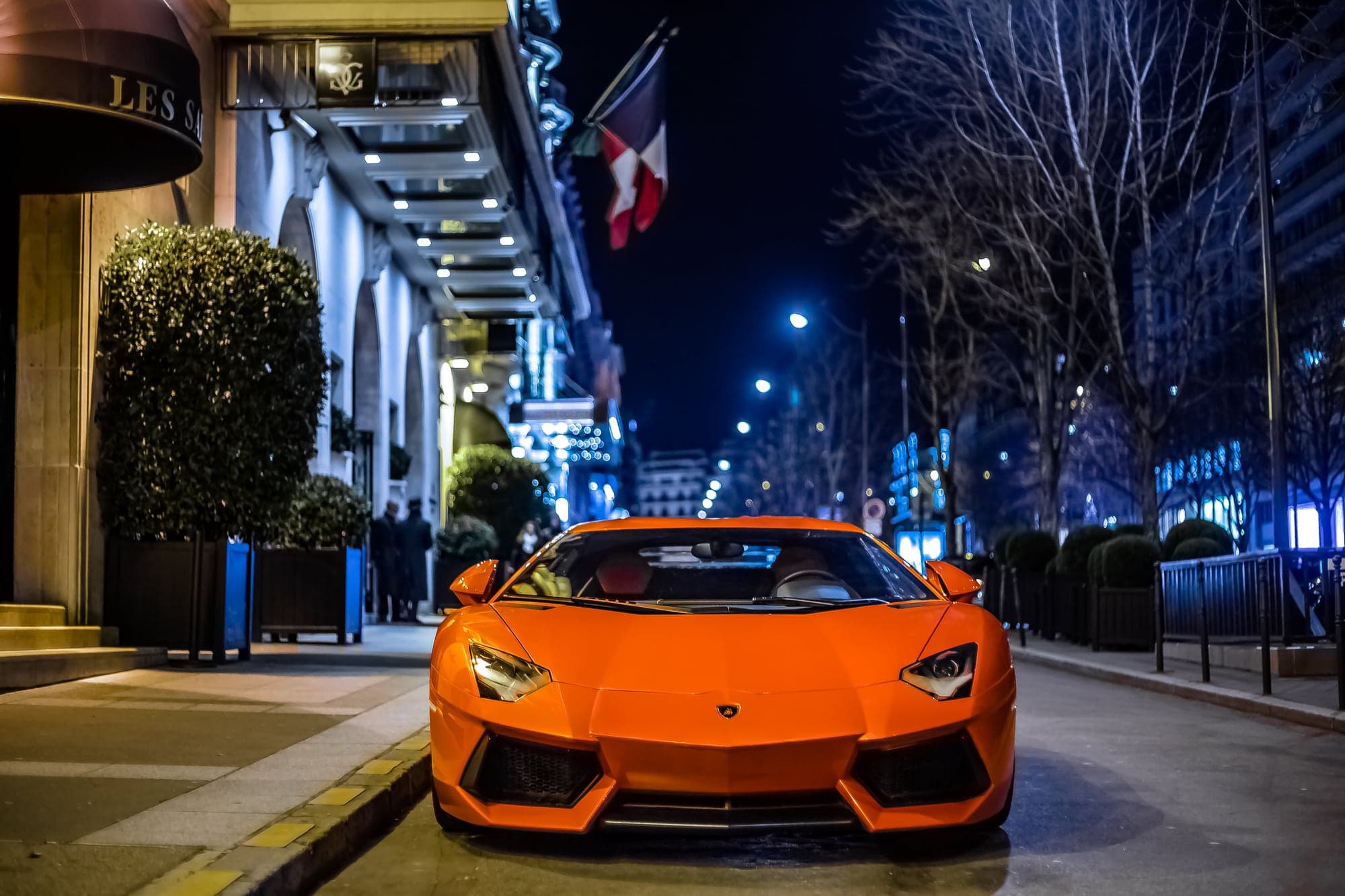Une Lamborghini Aventador orange garée de nuit devant l'entrée illuminée de l'hôtel George-V, à Paris, sous un drapeau français, avec des arbres dénudés et des réverbères en arrière-plan
