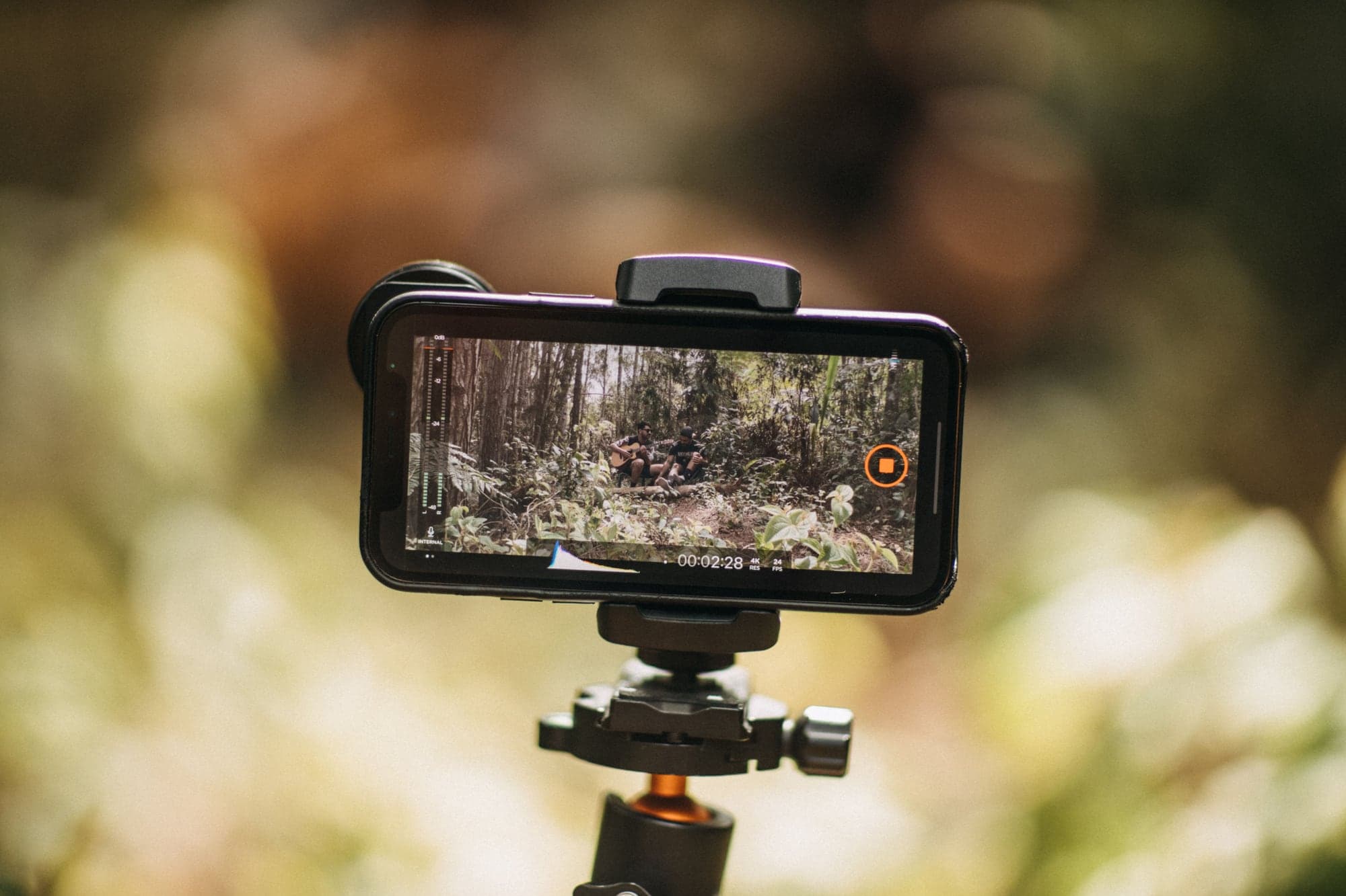 Smartphone fixé à l'horizontale sur la rotule d'un trépied, en extérieur, pendant un enregistrement : l'écran montre deux musiciens dans une forêt, un compteur à 00:02:28 et les indications 4K et 24 FPS