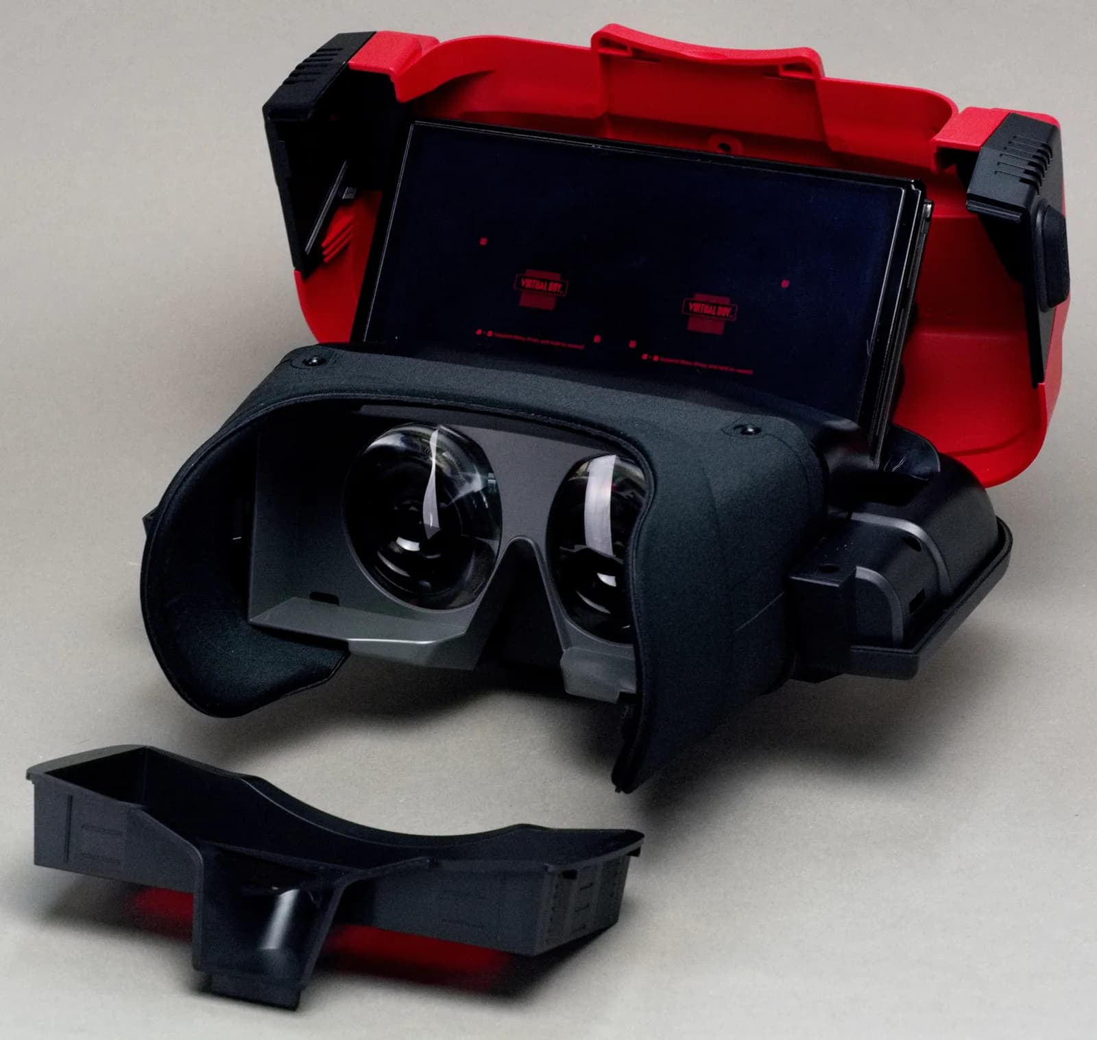 L'accessoire Virtual Boy pour Nintendo Switch, coque rouge ouverte, avec une Nintendo Switch OLED glissée à l'intérieur qui affiche l'écran de démarrage Virtual Boy derrière les deux lentilles de l'oculaire