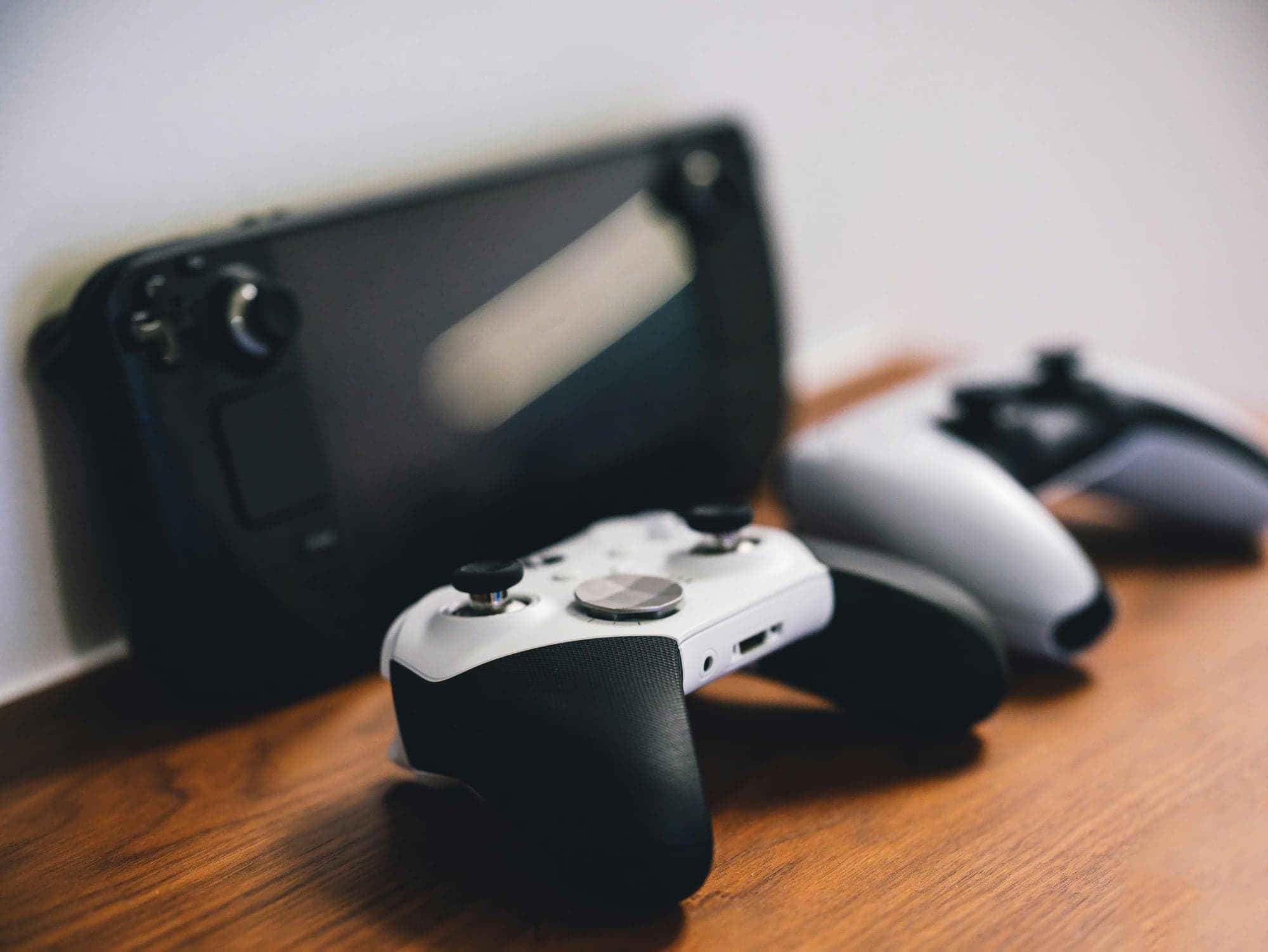 Une manette Xbox Elite Series 2 blanche au premier plan et une manette DualSense de PS5 posées sur un meuble en bois, devant une console portable Steam Deck