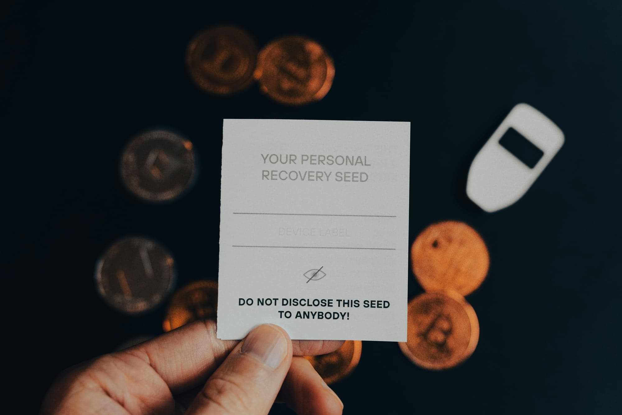 Une main tient une carte de sauvegarde vierge intitulée « Your personal recovery seed » et portant l'avertissement « Do not disclose this seed to anybody! », devant des pièces de cryptomonnaies et un portefeuille matériel blanc posés sur un fond sombre