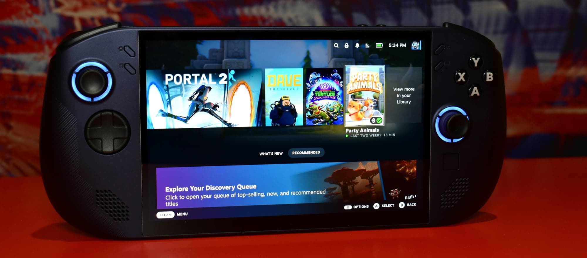 Console portable PC de Lenovo posée sur une table rouge, écran allumé sur l'accueil de Steam : Portal 2, Dave the Diver, Teenage Mutant Ninja Turtles: Splintered Fate et Party Animals