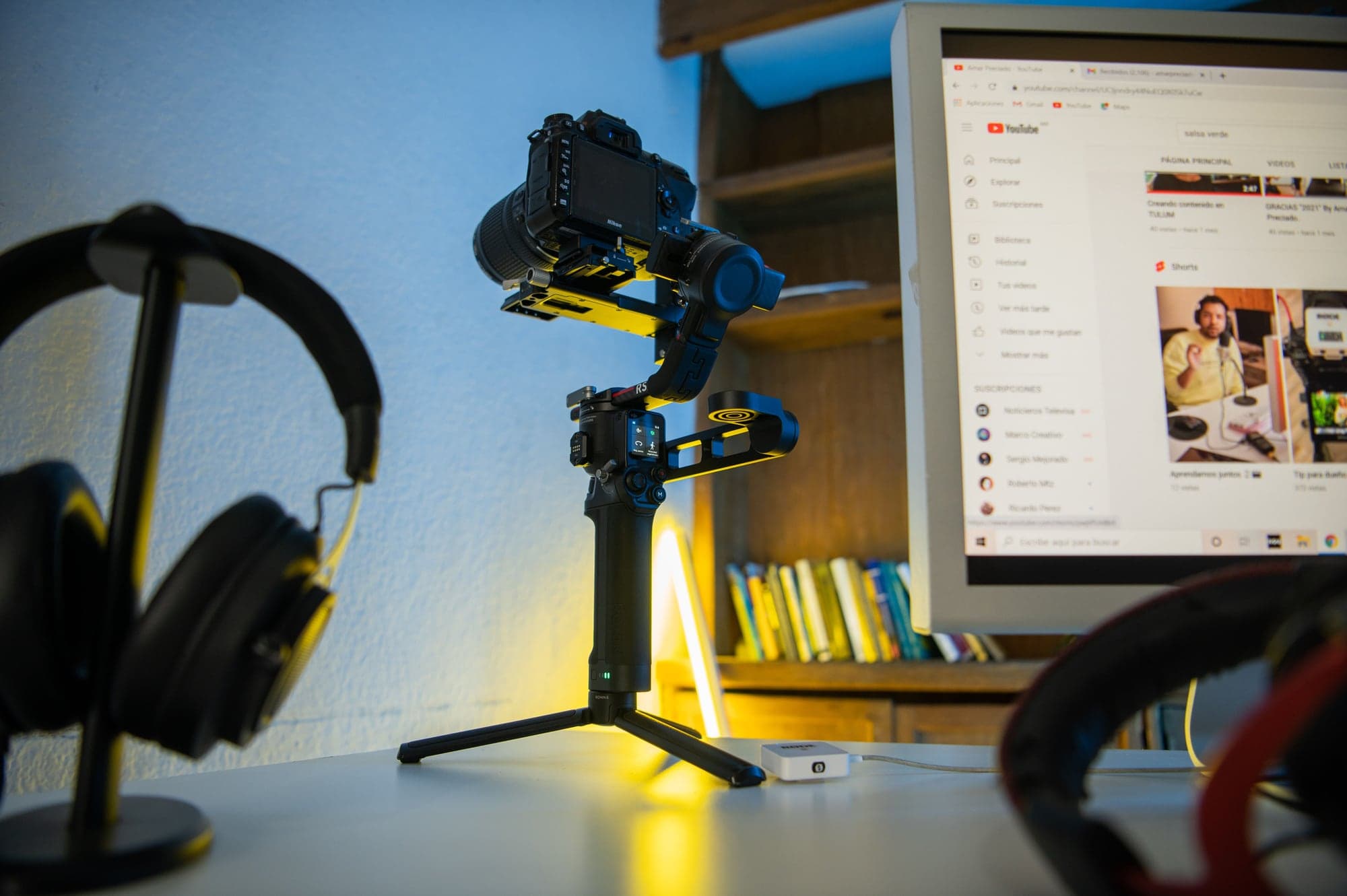 Appareil photo reflex Nikon monté sur un stabilisateur motorisé, posé sur un bureau entre un casque audio et un écran qui affiche la page d'une chaîne YouTube