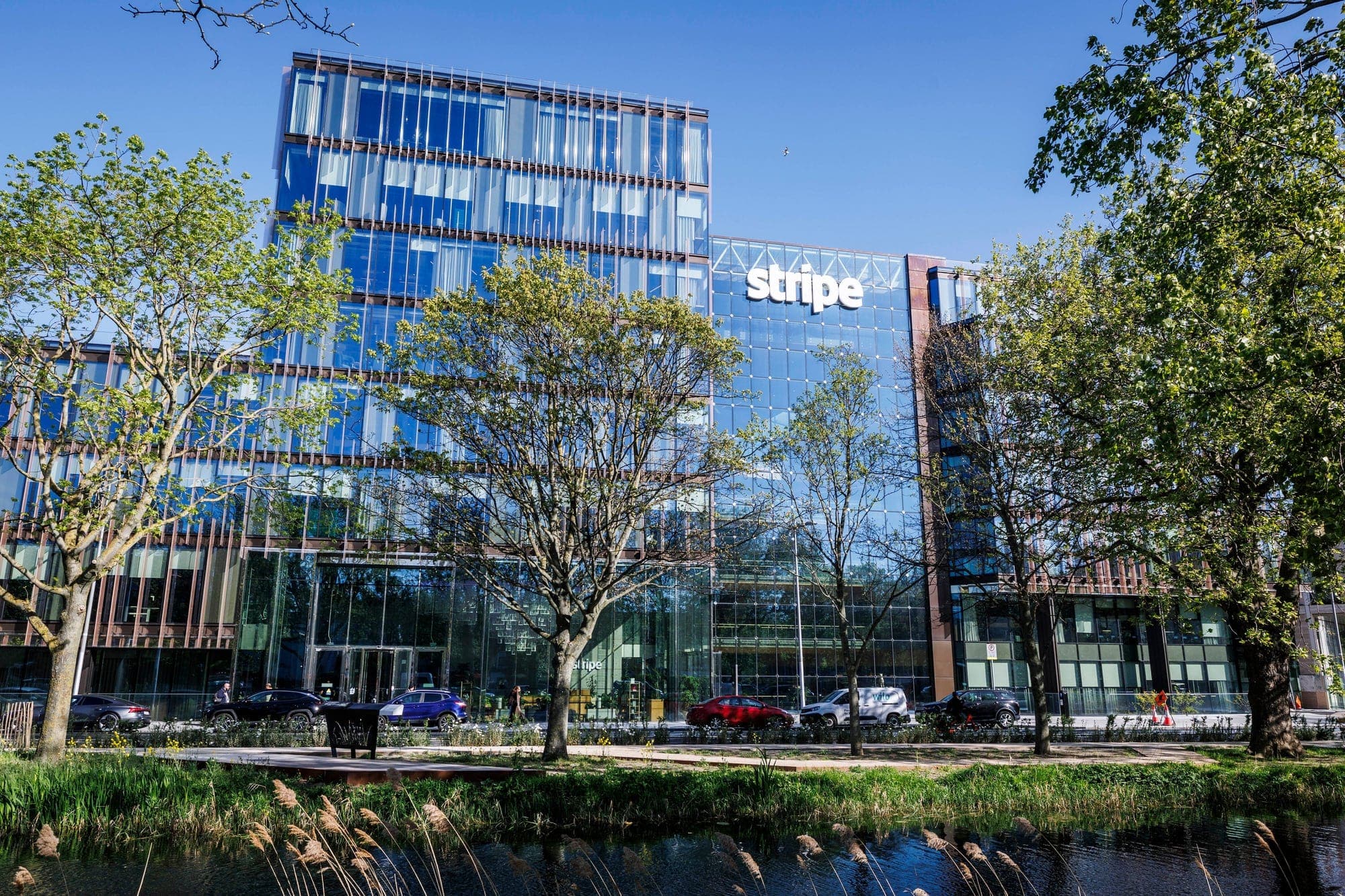 Façade vitrée des bureaux de Stripe à Dublin, avec le logo « stripe » en lettres blanches, derrière une rangée d'arbres au bord d'un canal