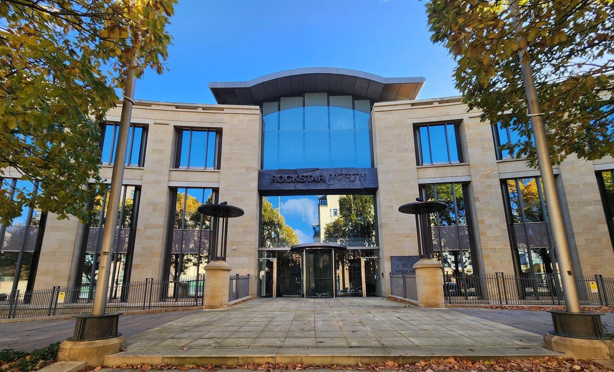 Façade en pierre claire et en verre du siège de Rockstar North à Édimbourg, avec l'enseigne du studio au-dessus de l'entrée, entre deux arbres aux feuilles d'automne