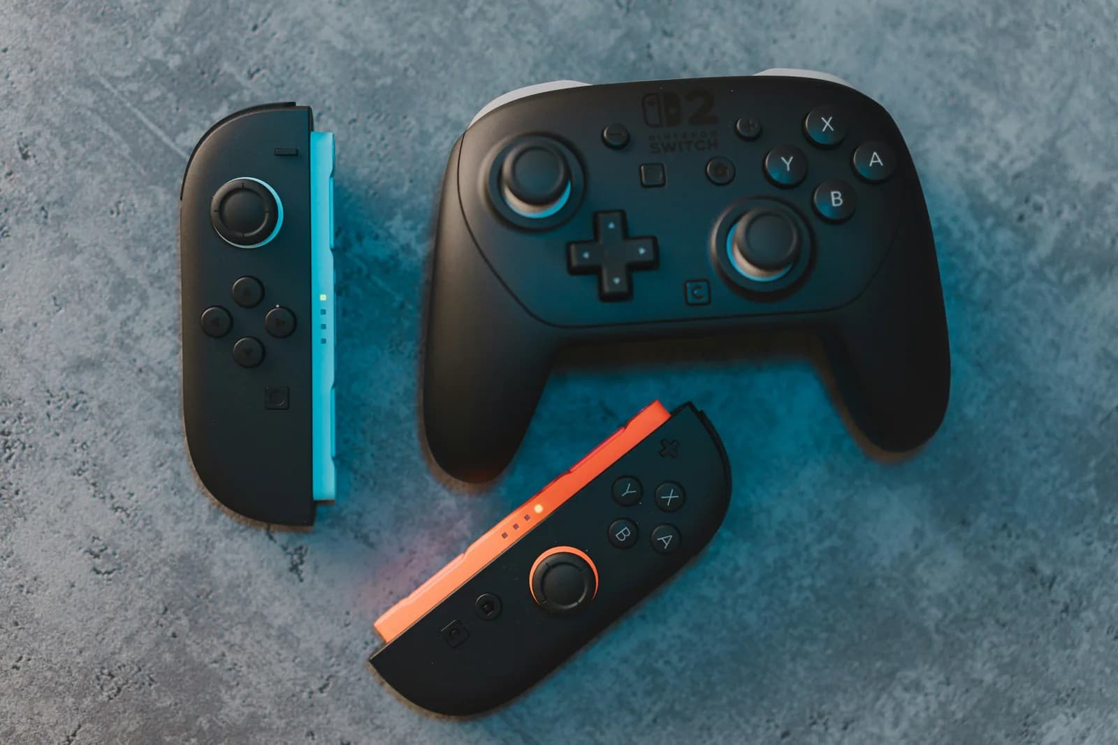 Un Joy-Con 2 gauche et un Joy-Con 2 droit, noirs à tranche bleue et orange, posés à côté d'une manette Pro Nintendo Switch 2 noire, sur une surface grise.
