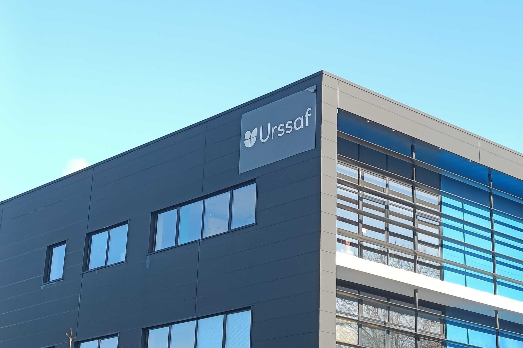 Angle d'un immeuble de bureaux gris et bleu portant l'enseigne « Urssaf », sous un ciel bleu, à Rennes