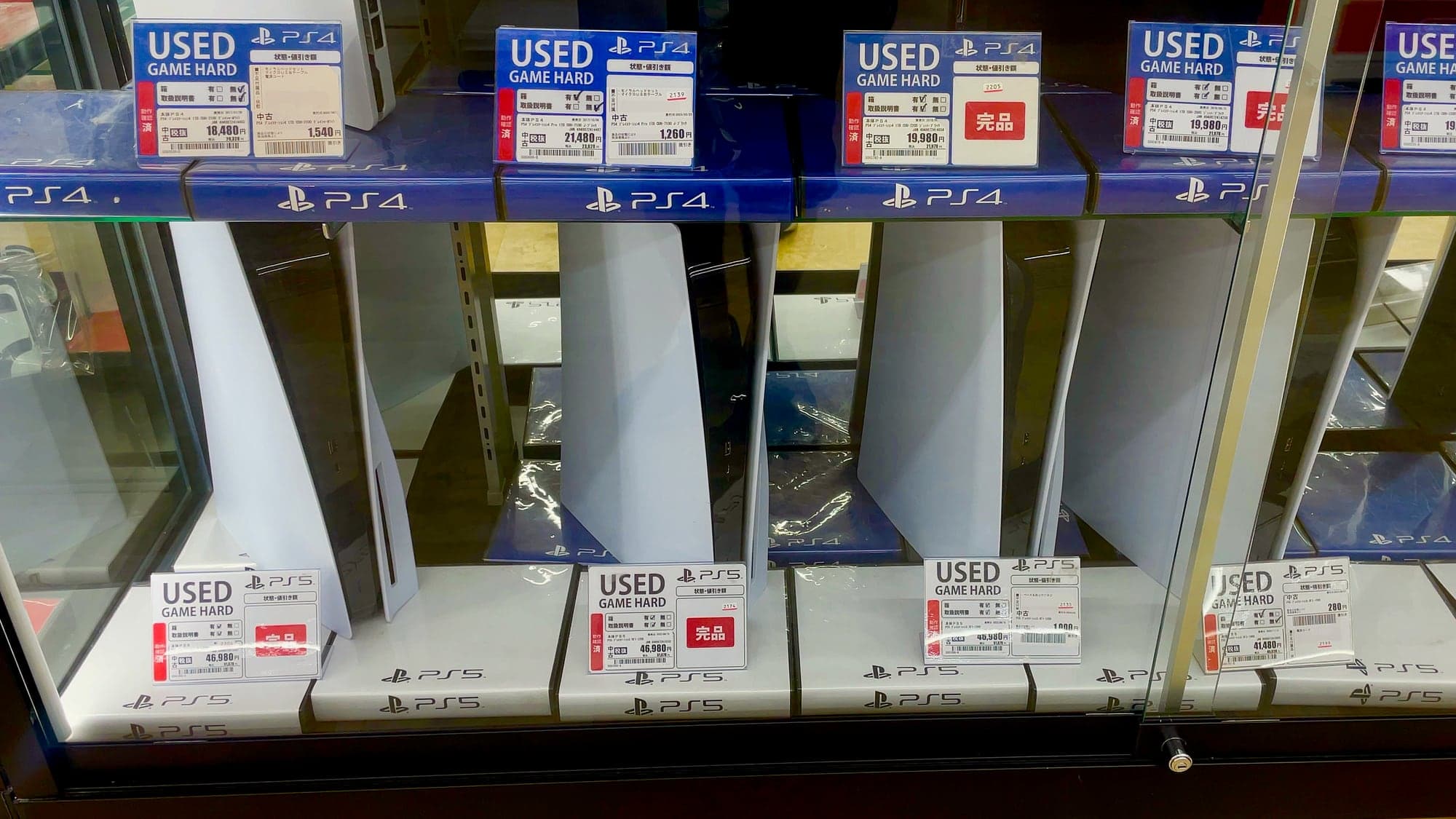 Vitrine d'une boutique de jeux vidéo au Japon : des PS5 blanches posées debout derrière une vitre, avec des étiquettes de prix « USED GAME HARD » en yens
