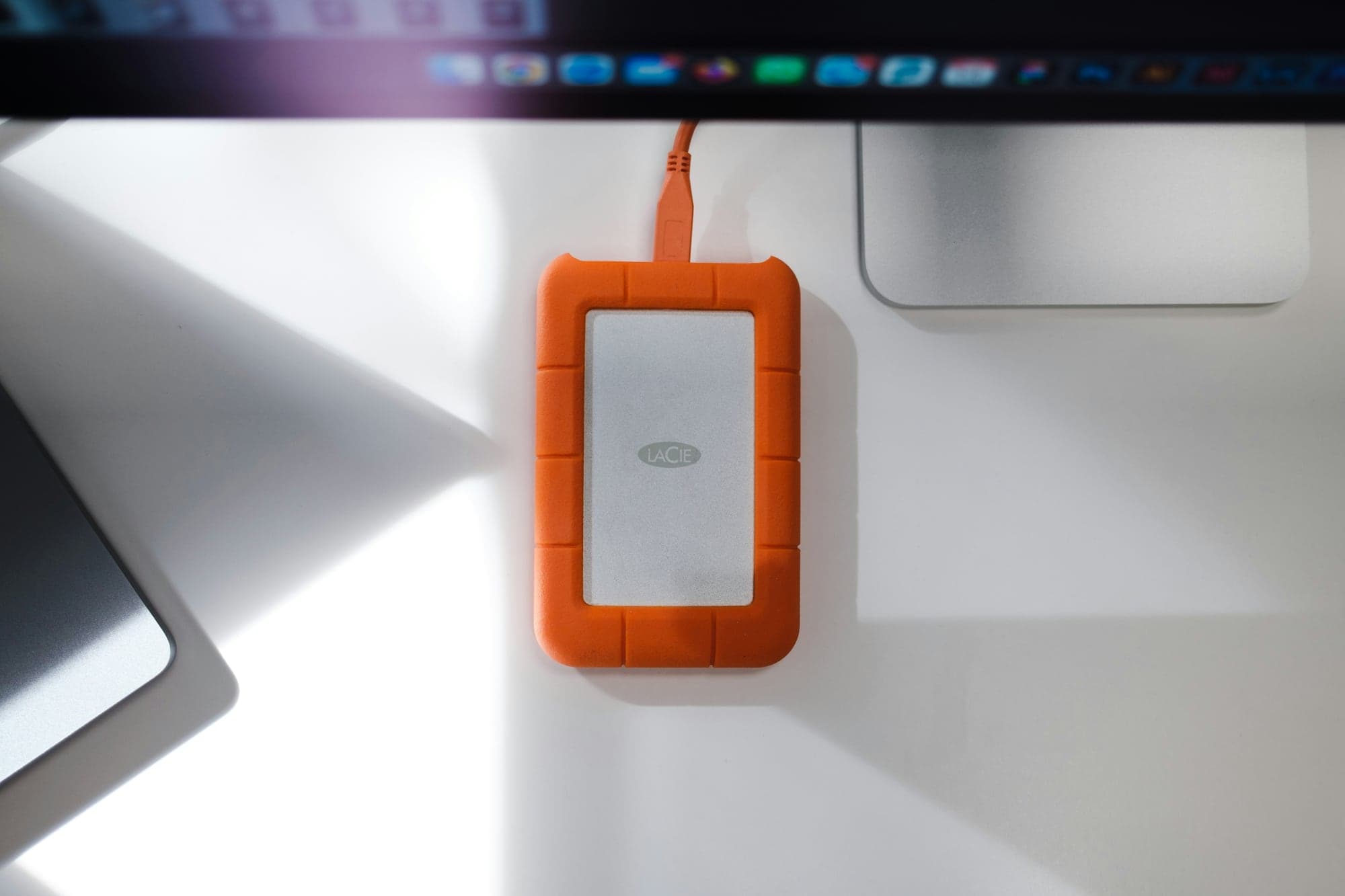 Disque dur externe LaCie Rugged à coque orange, relié par un câble USB orange et posé sur un bureau blanc, sous un écran d'ordinateur