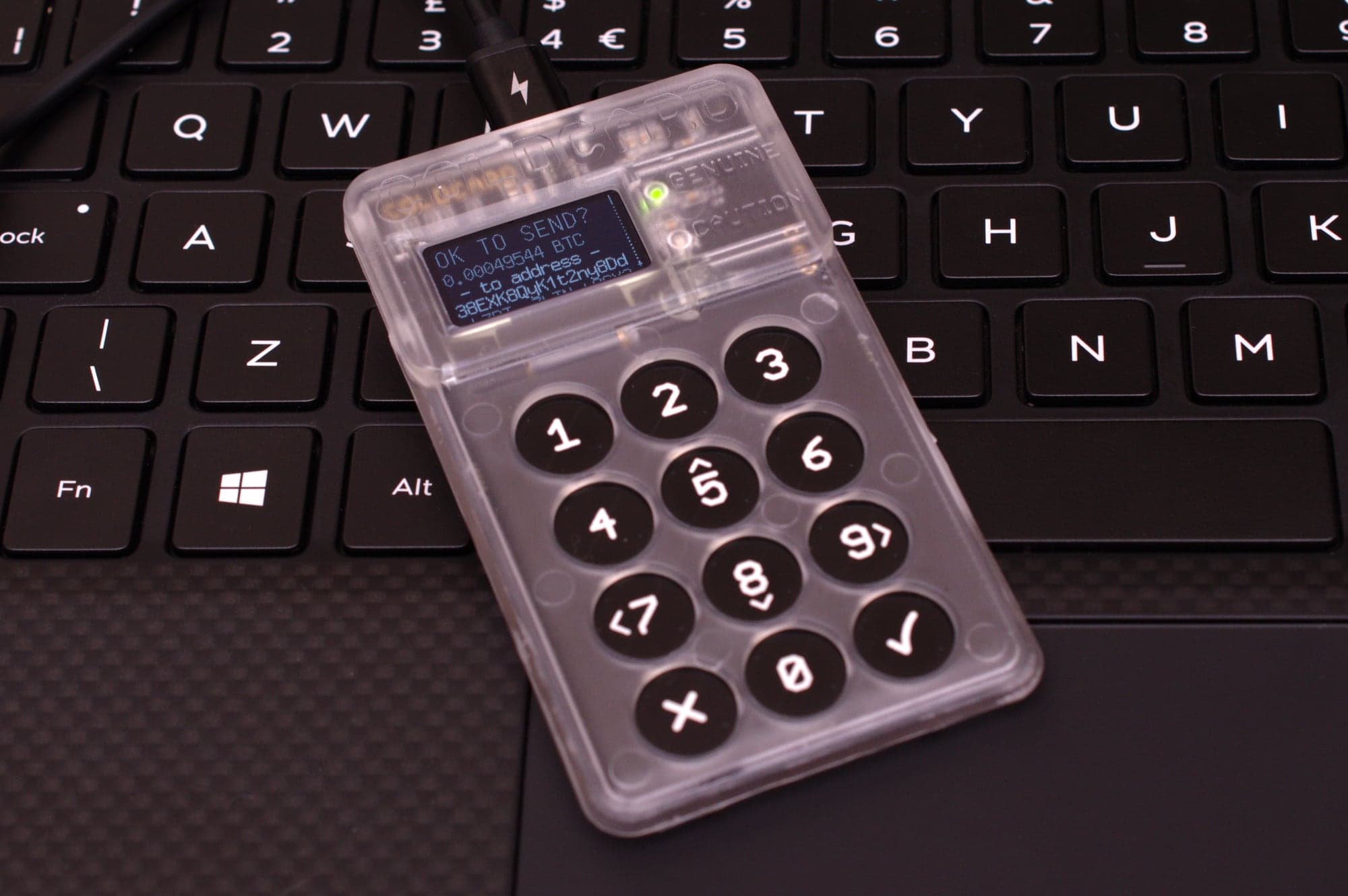 Portefeuille matériel Coldcard au boîtier transparent posé sur le clavier d'un ordinateur portable ; son écran demande « OK to send? » et affiche le montant en bitcoin et l'adresse de destination