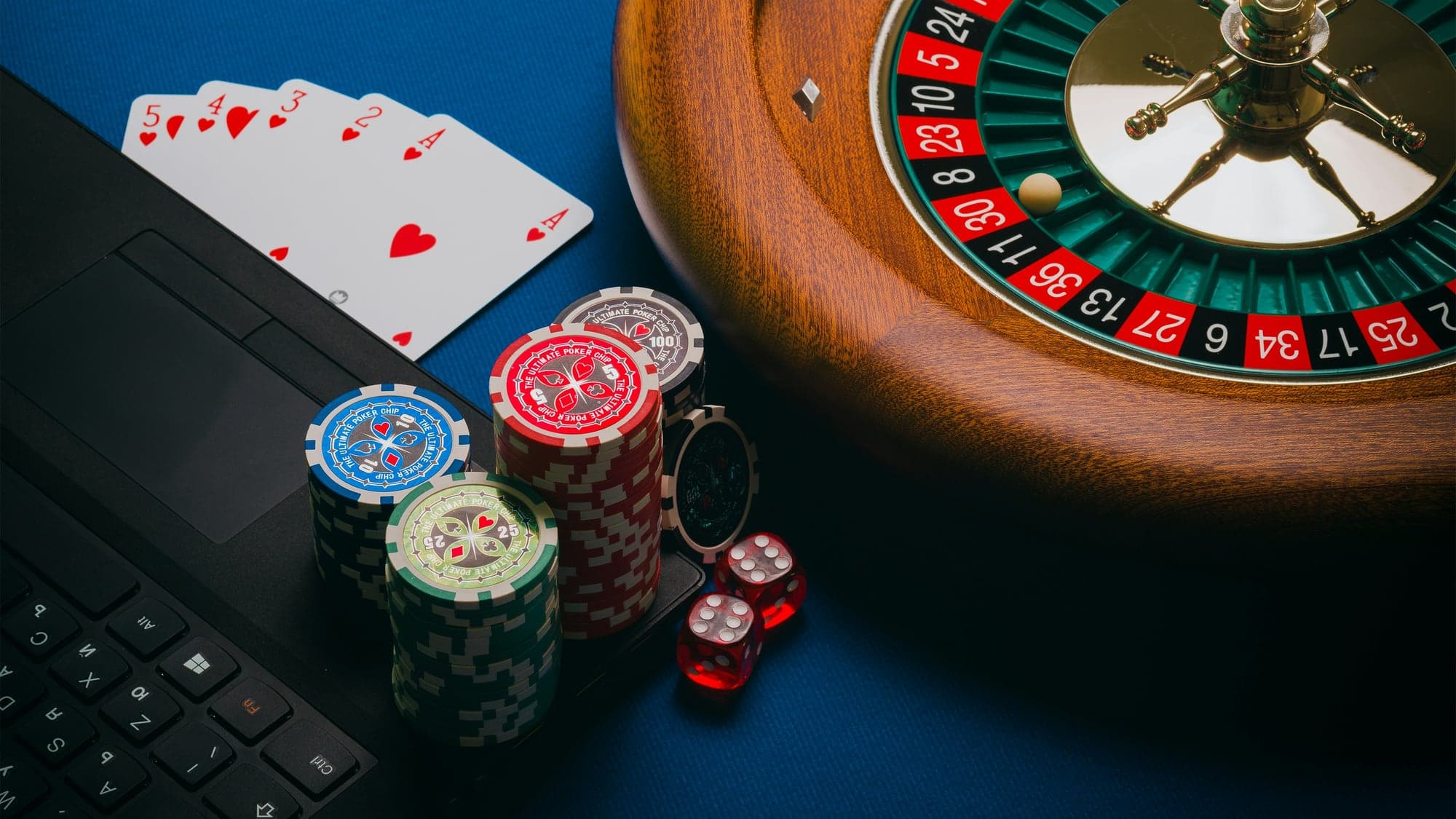 Jetons de poker empilés, deux dés rouges et des cartes à jouer posés sur un ordinateur portable, à côté d'un cylindre de roulette, sur un tapis bleu.
