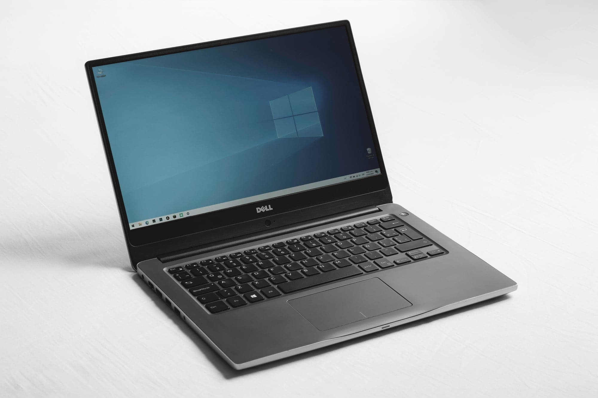 Ordinateur portable Dell ouvert sur une surface blanche, l'écran affichant le bureau de Windows 10 avec son fond bleu au logo Windows et sa barre des tâches